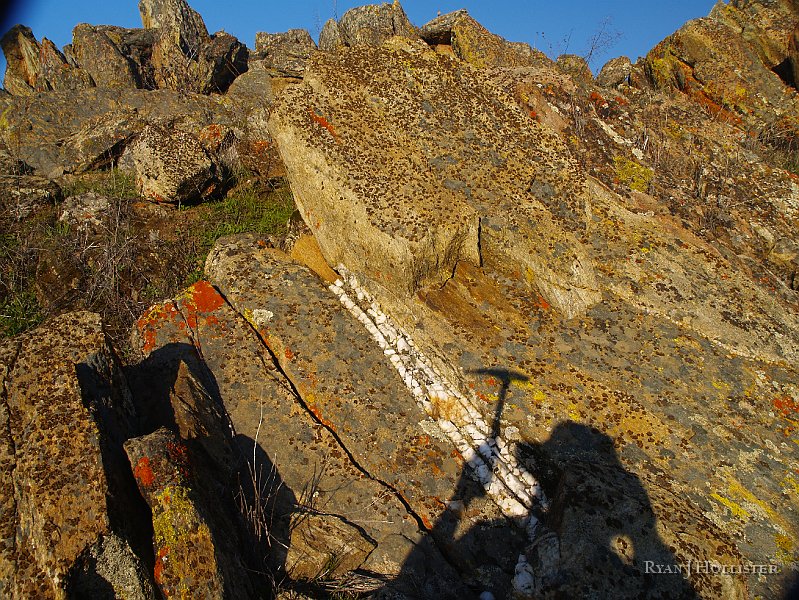 _1316498.JPG - Geology time-out:  The principle of cross-cutting relationships is very evident here.  The quartz vein is younger than the meta-sedimentary rocks that it intrudes.  The rock hammer shadow is for scale.  The upheaval of the rocks from horizontal left cracks in the rock into which the hydrothermal fluids intruded.